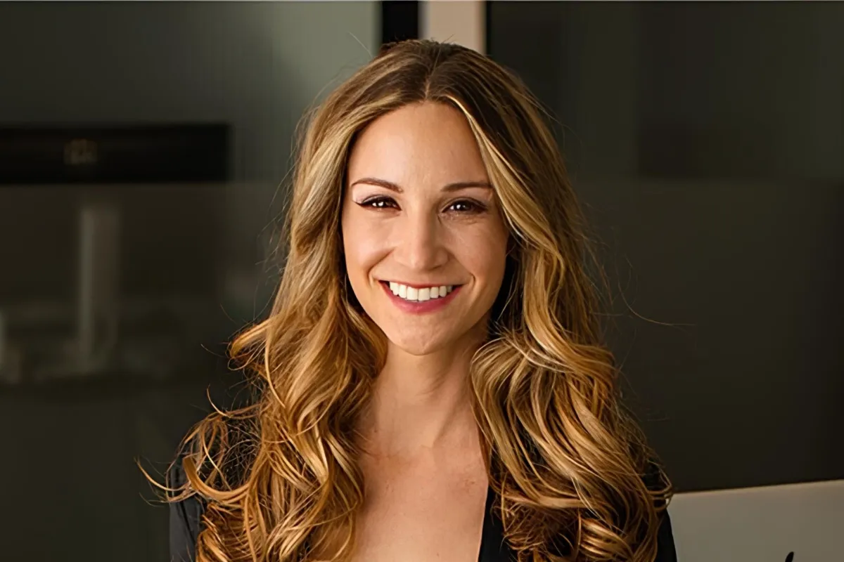 Smiling woman with long, wavy blonde hair and a black top, standing in a modern office setting.