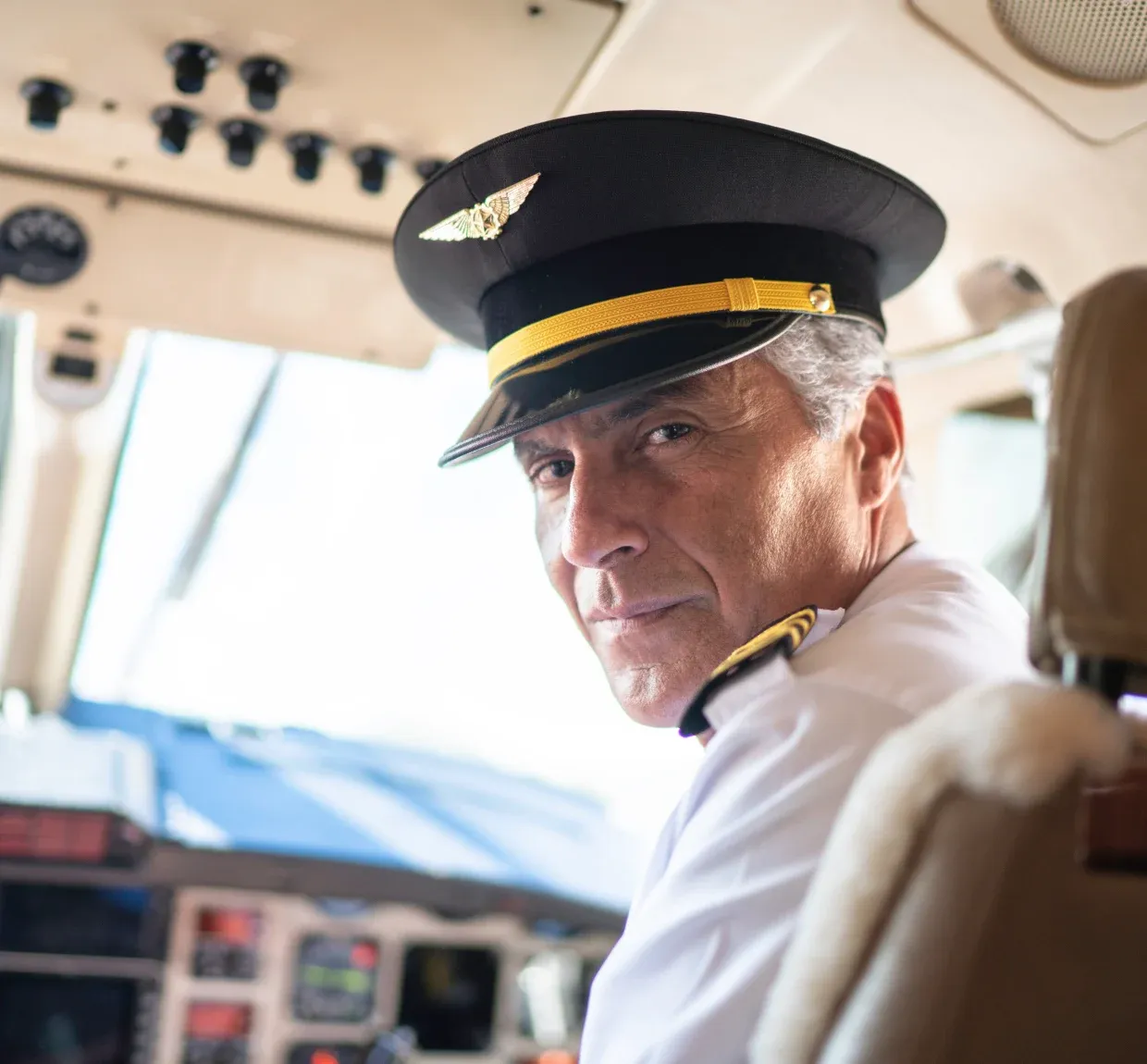 Pilot in uniform with a cap sits in an airplane cockpit, looking back over his shoulder, with control panels visible in the background.