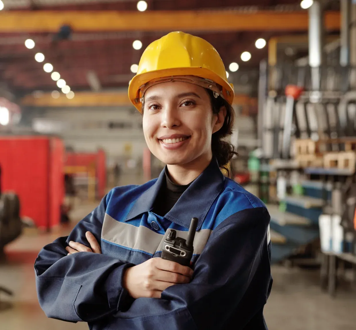 Smiling worker in a yellow hard hat and blue uniform holds a walkie-talkie in an industrial setting.