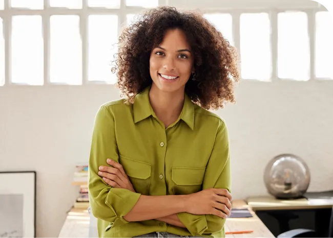 Smiling woman with curly hair in a green shirt, arms crossed, stands in a bright office with books and a globe on the desk.