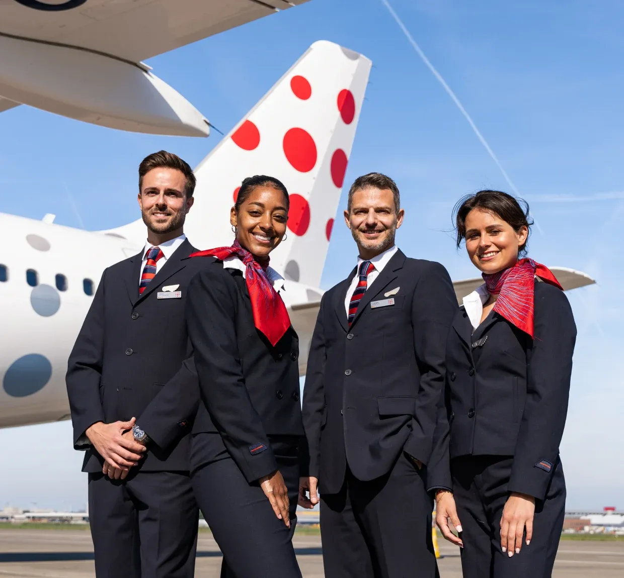 Four flight attendants in uniform stand smiling in front of a white airplane with red polka dots on the tail, under a clear blue sky.