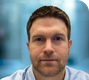 A man with short brown hair and a beard, wearing a light blue shirt, looks directly at the camera with a blurred background.