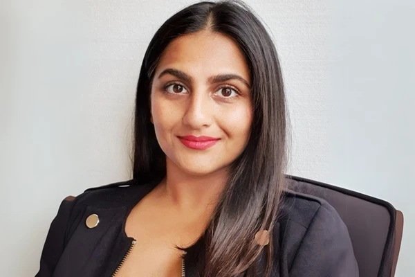 A woman with long dark hair and a black jacket smiles softly, sitting against a plain white background.