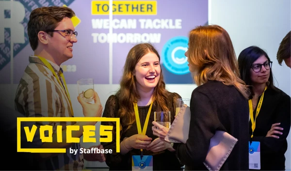 People networking at a conference event, wearing name tags and holding drinks. A "Voices by Staffbase" sign is visible in the foreground.