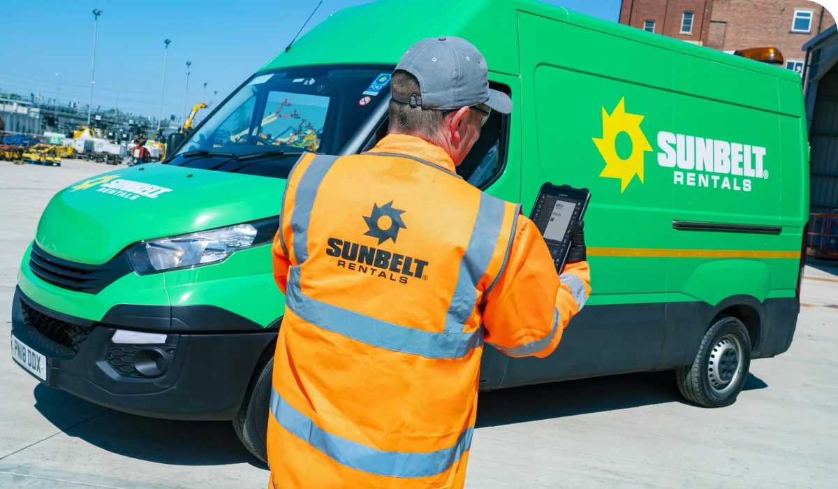 Worker in an orange Sunbelt Rentals jacket uses a tablet near a green Sunbelt Rentals van in an industrial area.