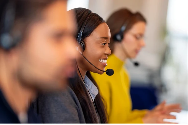 Three customer service representatives wearing headsets, focused and smiling, working at desks in a bright office environment.