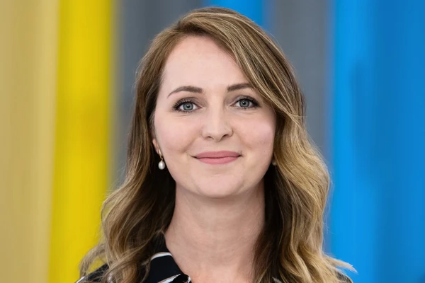 Smiling woman with wavy hair and pearl earrings, standing against a colorful background with yellow, gray, and blue panels.