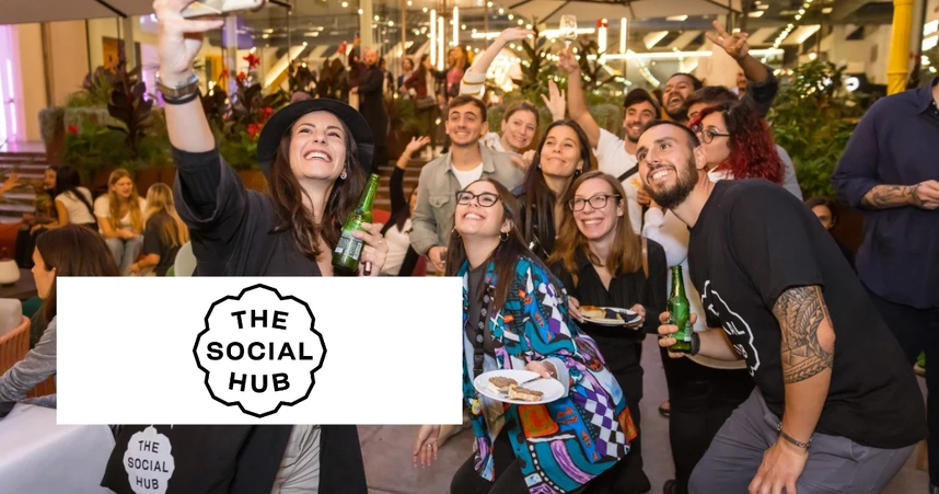 A group of people smiling and holding drinks at a lively indoor event, with "The Social Hub" logo in the foreground.