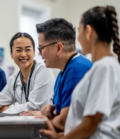 Three healthcare professionals in scrubs and a lab coat are smiling and conversing at a table in a medical setting.