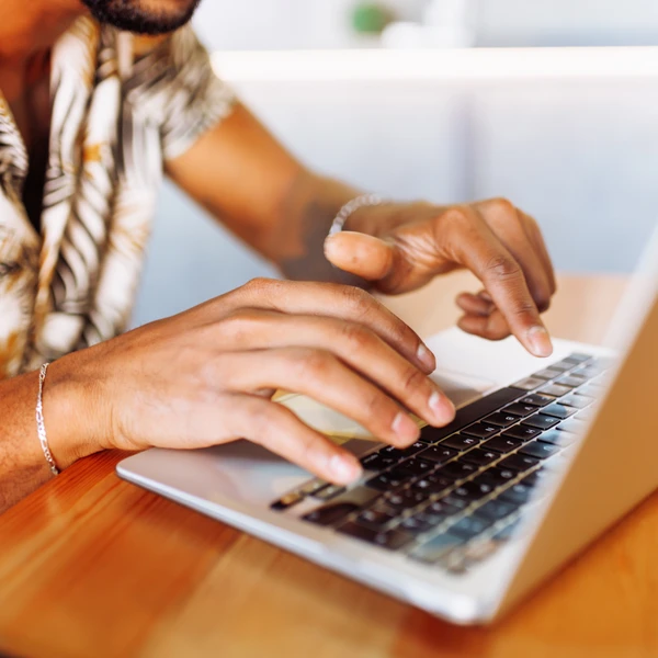 Person typing on a laptop at a wooden table, wearing a floral shirt and a bracelet, focusing on the keyboard.