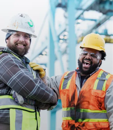 Two construction workers in safety gear smiling and laughing, with a blurred industrial background. One wears a white helmet, the other a yellow one.