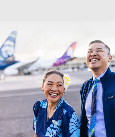 Two airline staff smiling on the tarmac, with airplanes in the background. The woman wears a flower in her hair.
