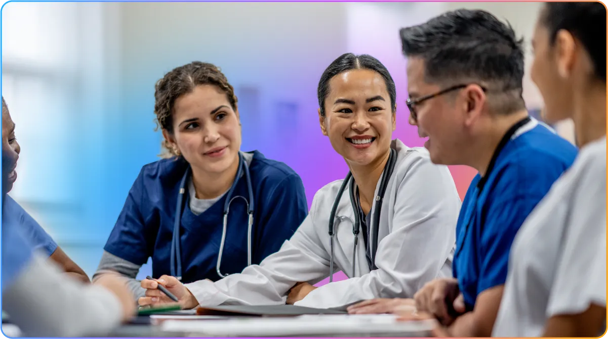 A diverse group of smiling healthcare professionals in scrubs and lab coats having a discussion around a table.