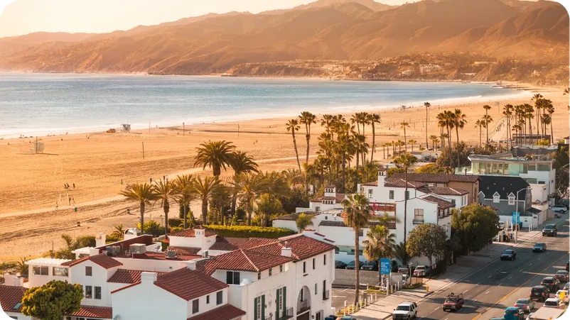 Aerial view of Santa Monica beach at sunset, with palm trees, coastal homes, and mountains in the background.