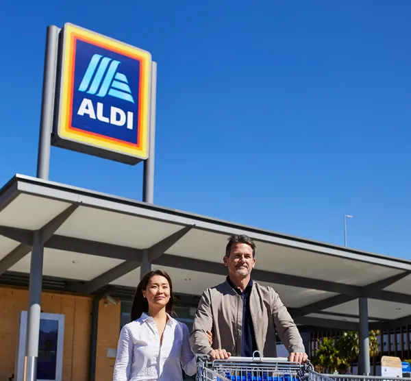 A man and woman stand with a shopping cart in front of an Aldi store under a clear blue sky.