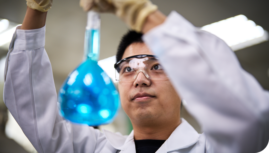 A scientist in a lab coat and goggles examines a blue liquid in a flask, holding it up to the light.