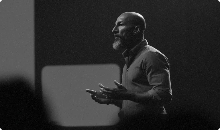 A bearded man speaks passionately on stage, gesturing with his hands. The background is blurred, creating a focused spotlight effect. Black and white.
