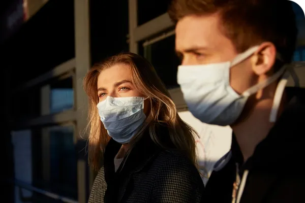 Two people wearing face masks stand outdoors in sunlight, looking in the same direction.