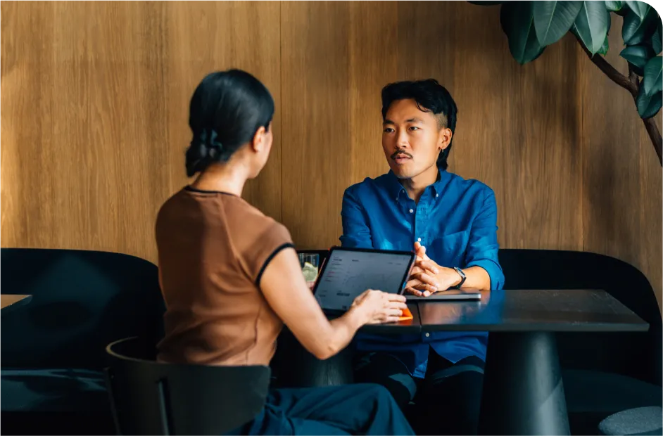 Two people sitting at a table in a cafe, engaged in conversation. One is using a laptop, and the setting has wooden walls and a large plant.