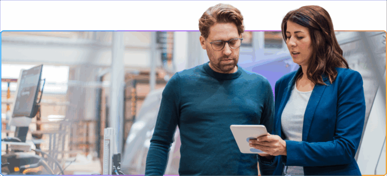 Two professionals standing in an industrial setting, looking at a tablet together during a discussion.