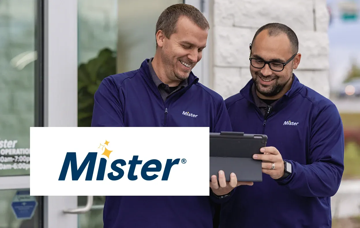 Mister logo over photo of two employees in branded jackets smiling while looking at a tablet.