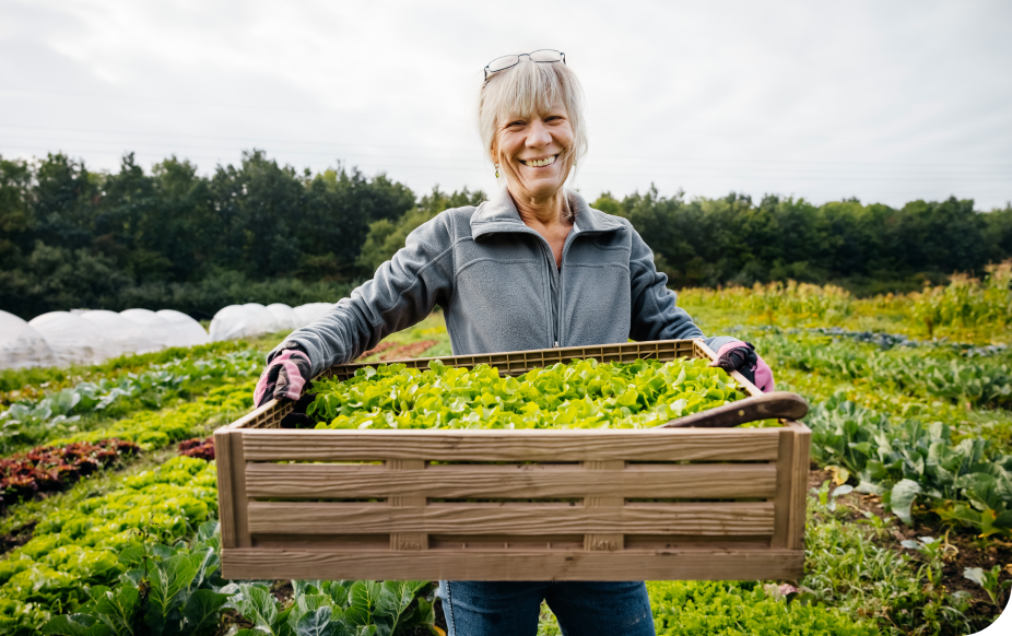 Smiling woman holding a wooden crate full of fresh greens in a lush vegetable garden, wearing gloves and a jacket.