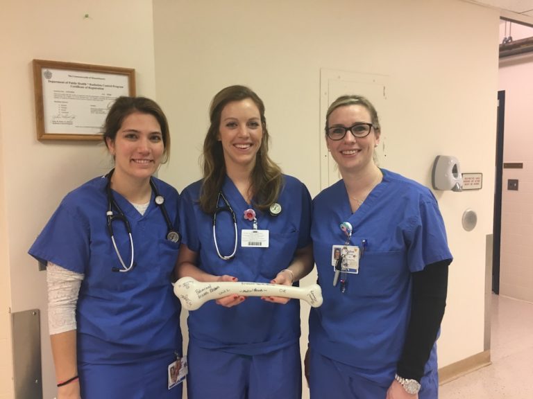 Three female healthcare professionals in blue scrubs smiling and standing together in a hospital hallway, holding a large model of a human bone.