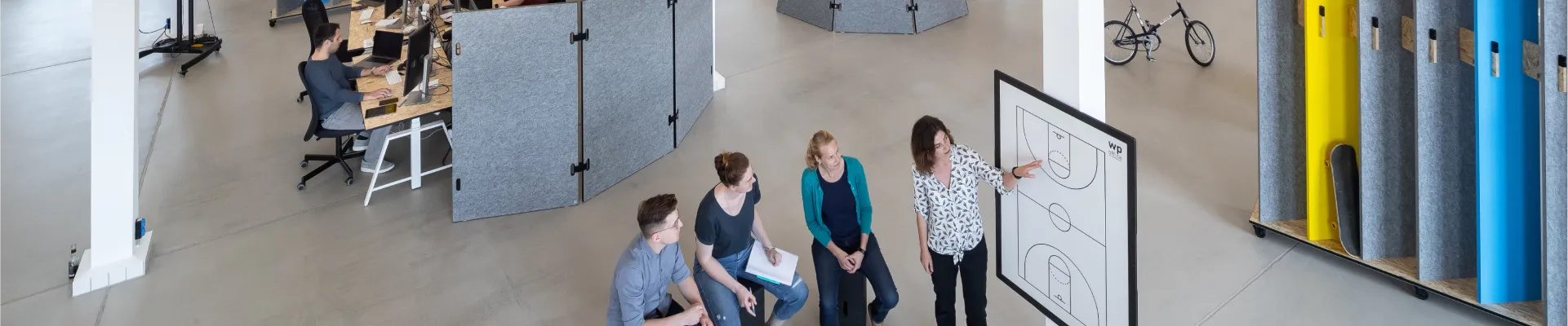 Open office space with people collaborating at a whiteboard. Desks with computers, a bicycle, and colorful dividers are visible.