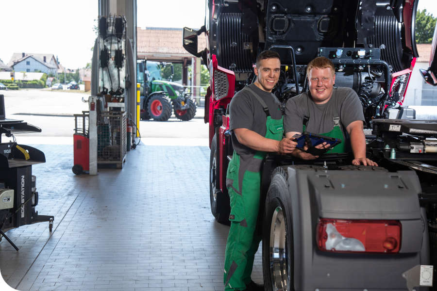 Two mechanics smiling while working on a truck in a garage, with tools around and a tractor visible outside.