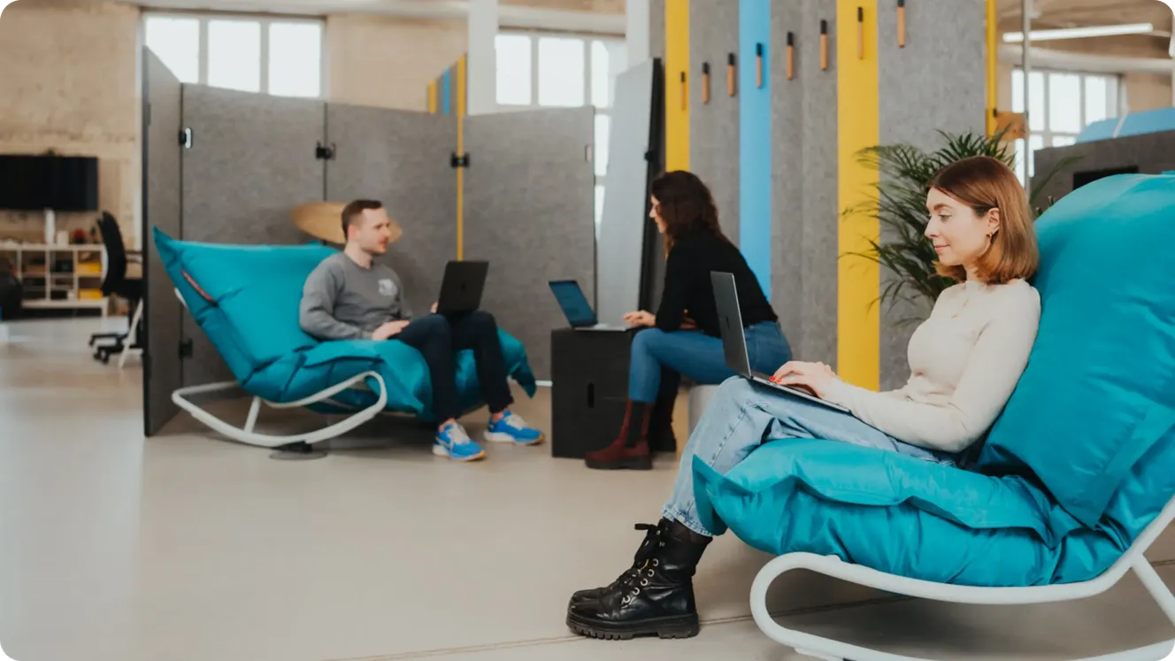 Staffbase colleagues working on laptops in teal lounge chairs in a colorful open office space.