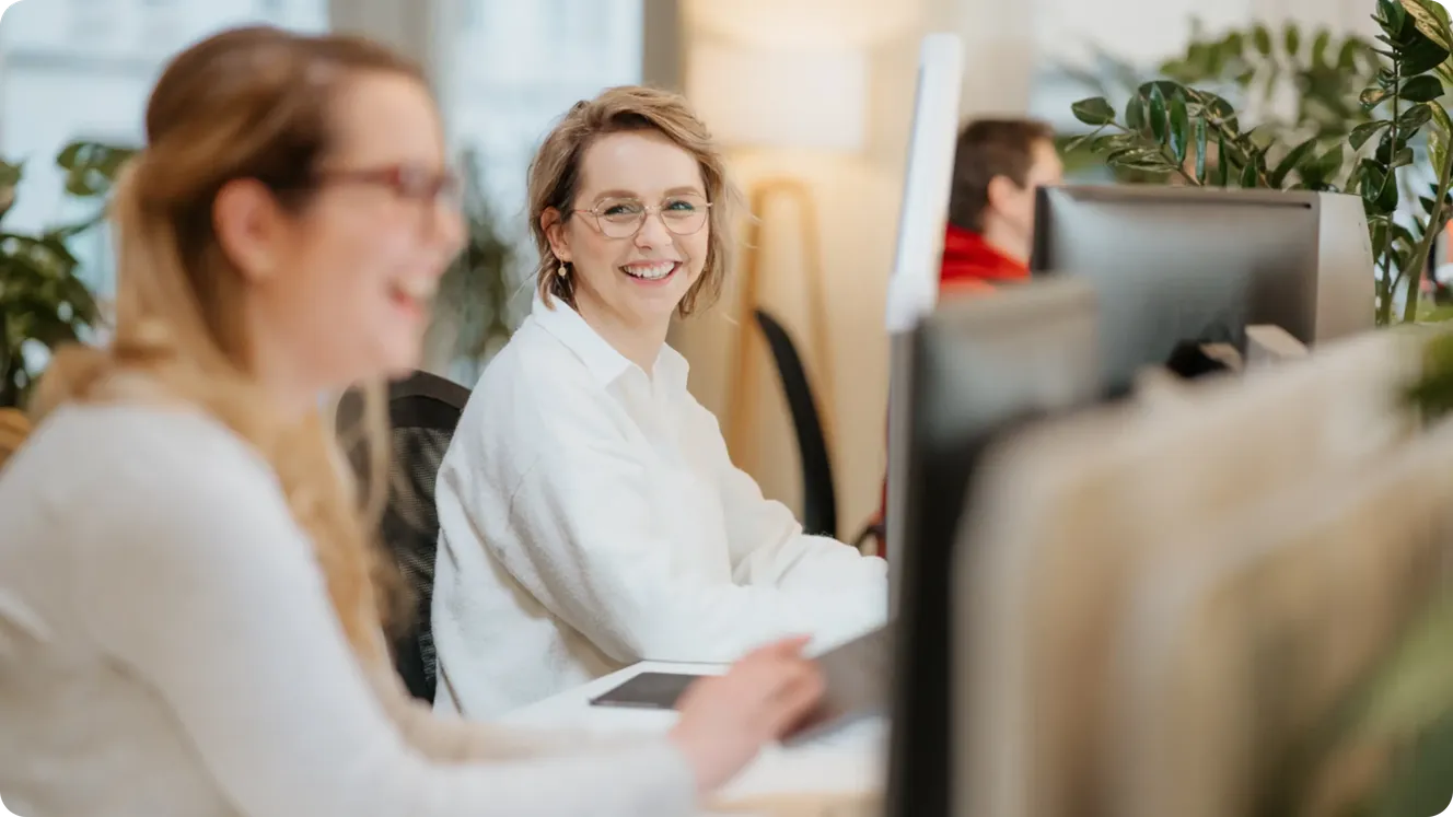 Two Staffbase colleagues smiling and chatting at their desks in an open-plan office with plants.