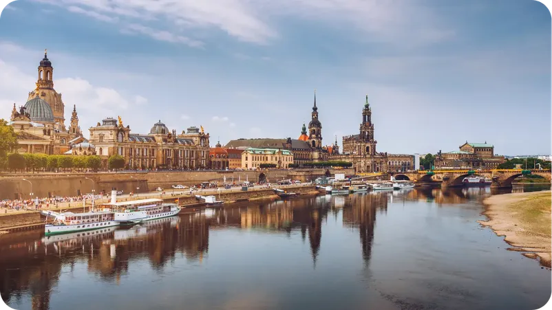 Dresden's historic skyline along the Elbe river, with domes, towers, and a river cruise boat docked.