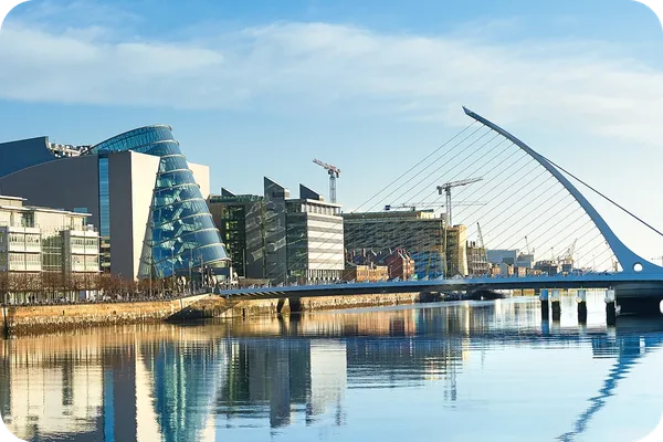 Modern cityscape with a river, glass buildings, cranes, and a distinctive white bridge reflecting in the calm water under a clear blue sky.