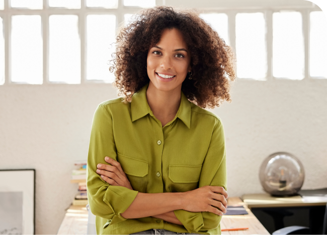 Smiling woman with curly hair in a green shirt, arms crossed, stands in a bright office with books and a globe on the desk.
