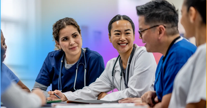 A diverse group of smiling healthcare professionals in scrubs and lab coats having a discussion around a table.