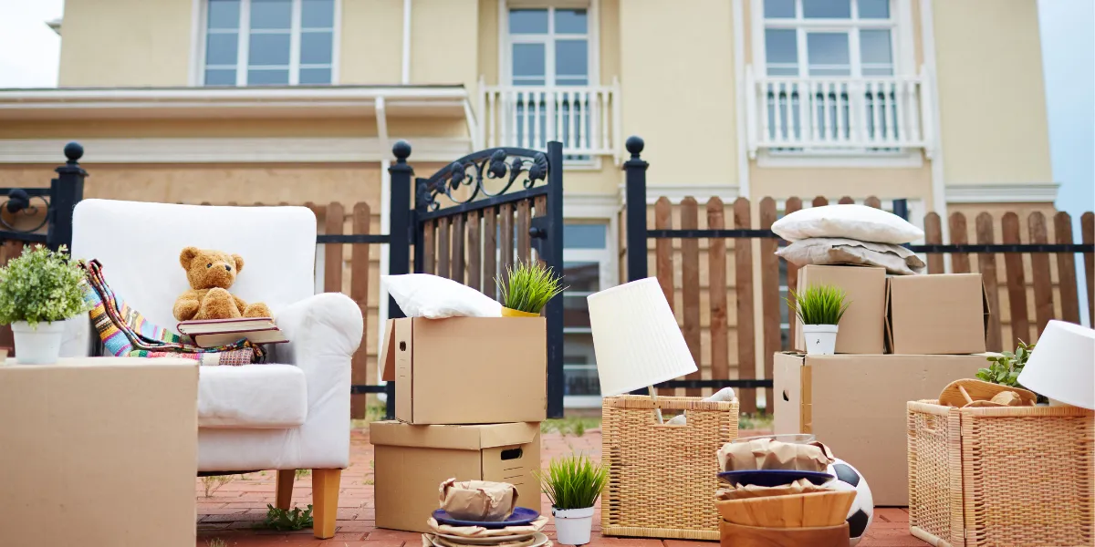 Moving day scene outside a house: a white armchair with a teddy bear and books, surrounded by cardboard boxes, wicker baskets, potted plants, a lamp, pillows, and packed household items on a brick patio.