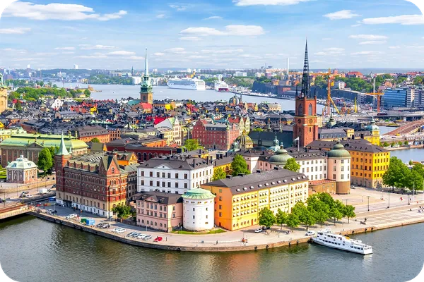 Aerial view of Stockholm's Gamla Stan with colorful historic buildings, water, boats, and a spire set against a partly cloudy sky.