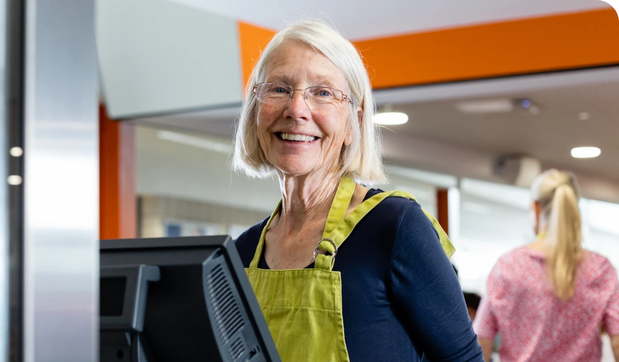 Smiling older woman with glasses and a green apron stands at a checkout counter, with a blurred person in the background.