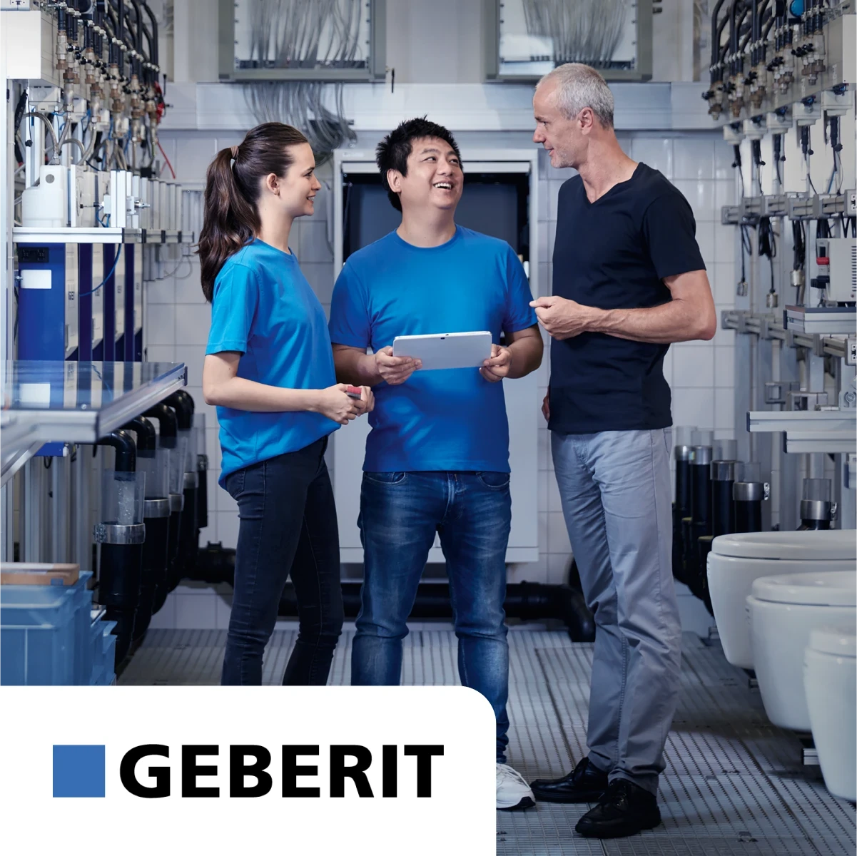 Three people in a plumbing showroom, two in blue shirts and one in black, standing and talking, with Geberit logo in the corner.