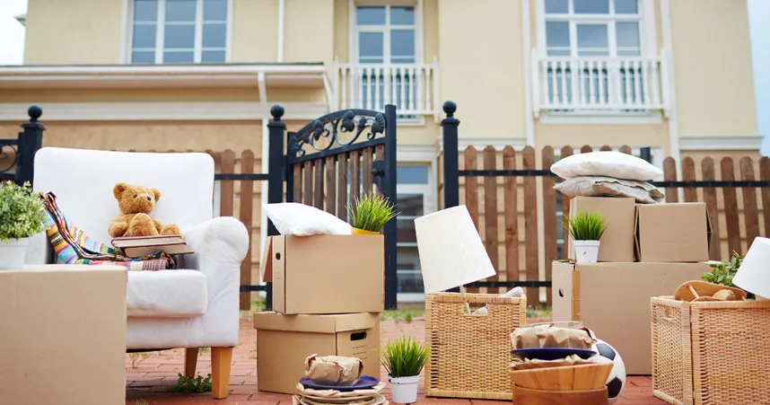 Moving day scene outside a house: a white armchair with a teddy bear and books, surrounded by cardboard boxes, wicker baskets, potted plants, a lamp, pillows, and packed household items on a brick patio.