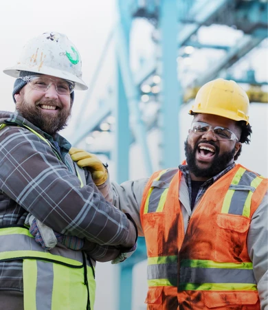 Two construction workers in safety gear smiling and laughing, with a blurred industrial background. One wears a white helmet, the other a yellow one.