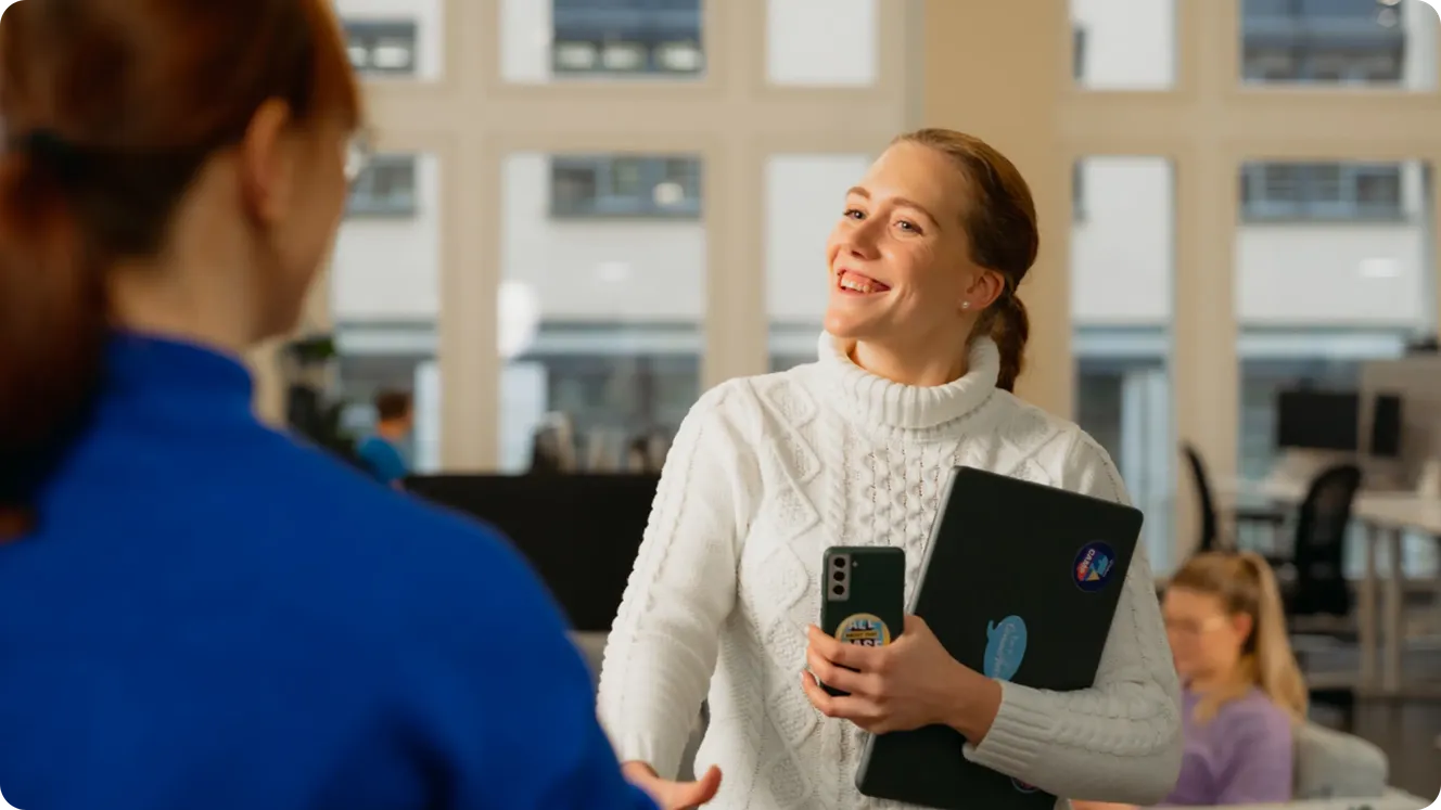 Staffbase employee holding a laptop and phone, smiling while greeting a colleague in the office.