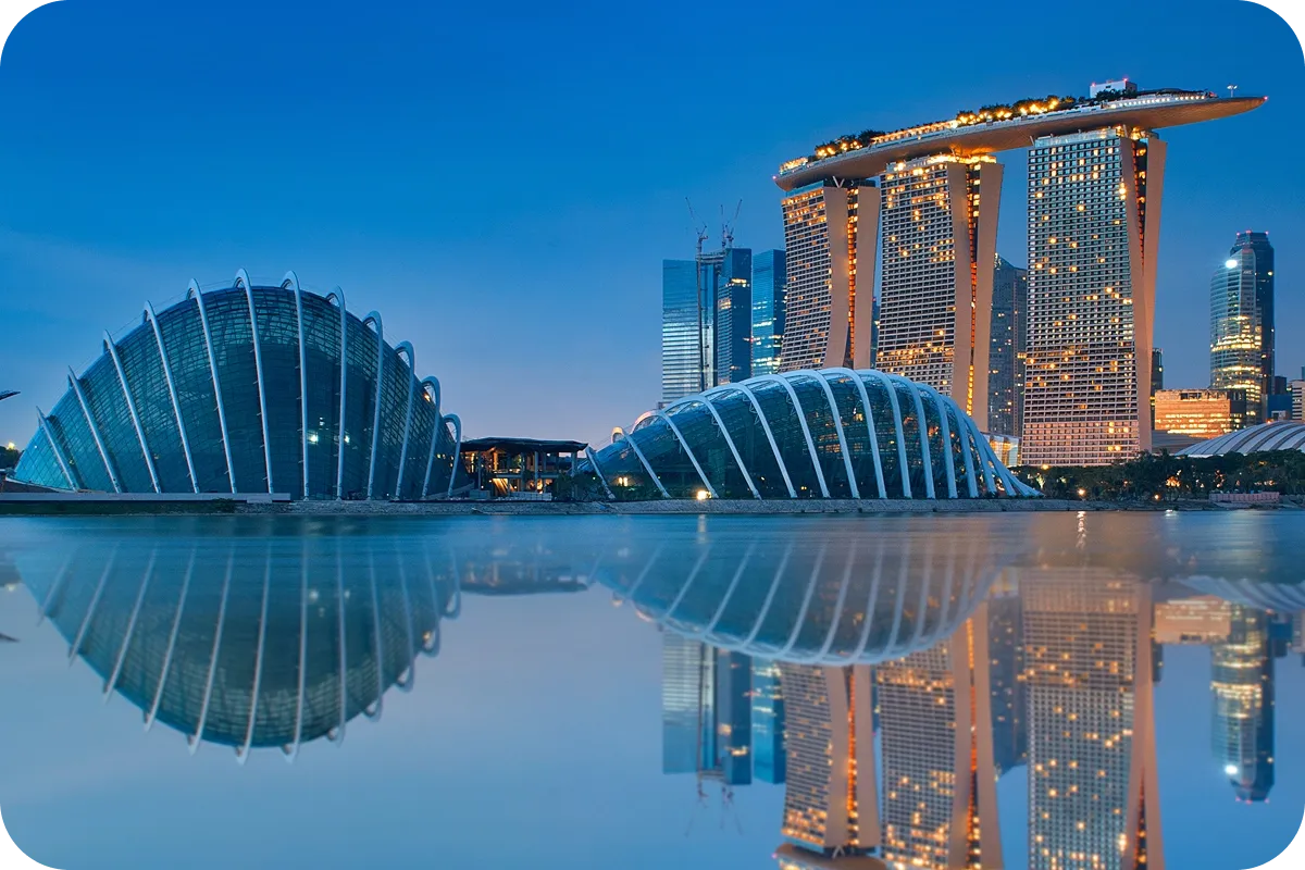 Skyline of Singapore at dusk, featuring Marina Bay Sands and futuristic domes, reflected in calm water under a clear blue sky.