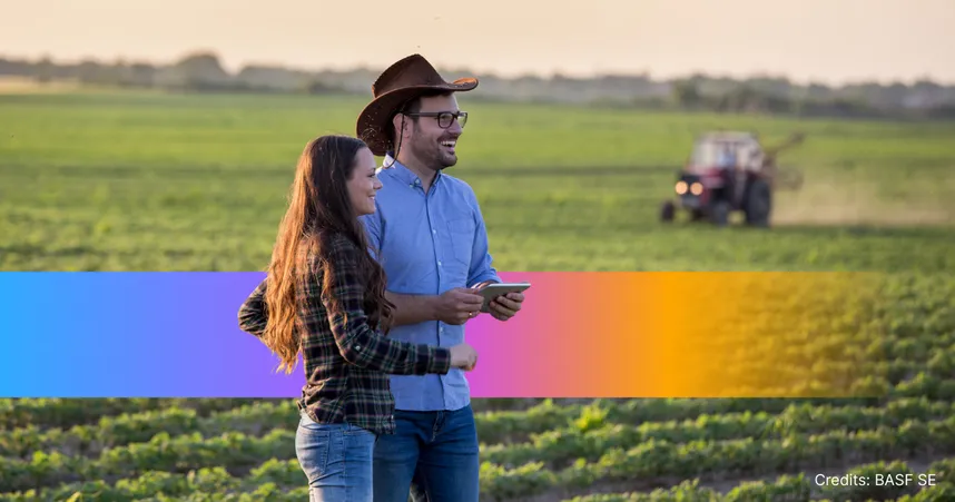 A man and woman stand together in a lush green field, smiling, with a tractor in the distance.
