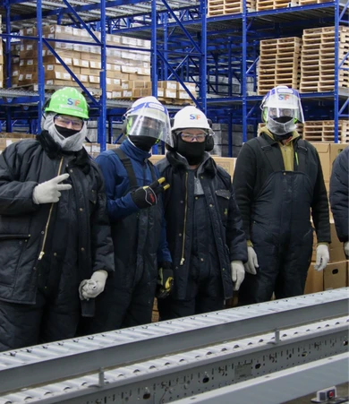 Four workers in protective gear and helmets stand in a warehouse with shelves of boxes in the background. Two of them are making peace signs.
