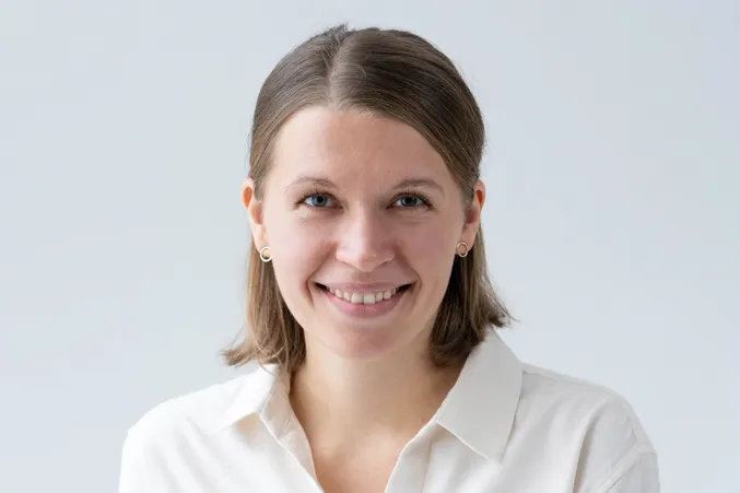 Smiling person with short brown hair, wearing a white shirt and small earrings, against a plain background.