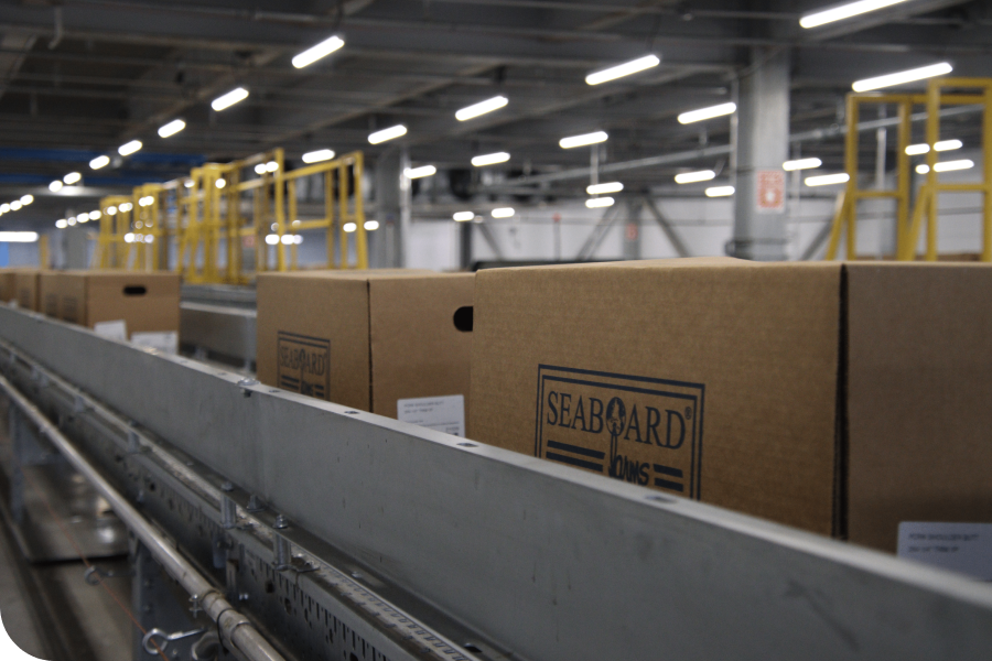 Conveyor belt with brown boxes labeled "SEABOARD" in an industrial warehouse with overhead lighting and yellow railings.
