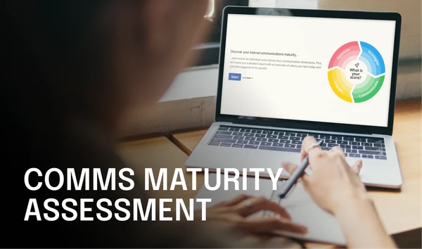 Person writing on paper with a laptop displaying a "Comms Maturity Assessment" diagram, featuring a colorful circular chart.