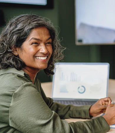 Smiling woman with gray hair sits at a table, a laptop displaying charts and graphs in the background.