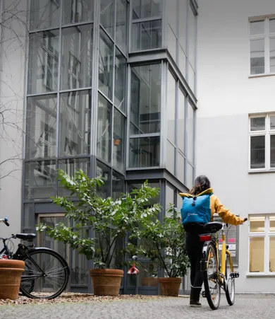 Staffbase employee with a branded bag arriving with her bike in a courtyard outside a glass-fronted office building, with another bike parked nearby.
